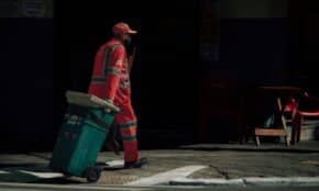 Municipal worker in uniform pulling a trash bin on an urban street.