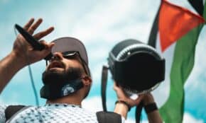 A man with a face mask speaks into a microphone at a peaceful protest holding a Palestinian flag.