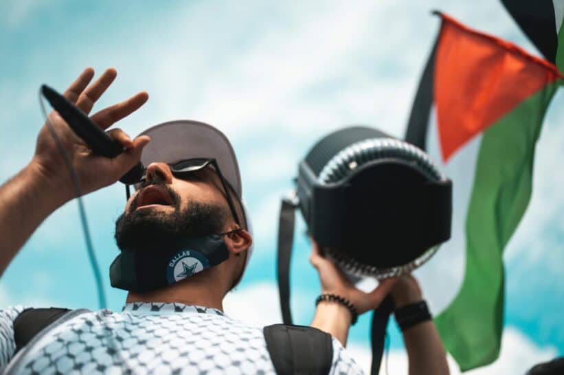 A man with a face mask speaks into a microphone at a peaceful protest holding a Palestinian flag.