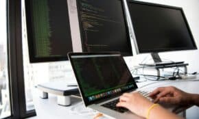 A developer writes code on a laptop in front of multiple monitors in an office setting.