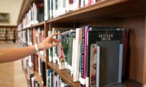A close-up of a person's hand reaching for a book in a library shelf, suggesting exploration and learning.