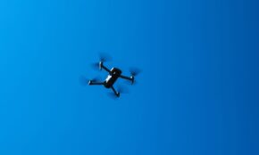 A black drone flying high in a clear blue sky, showcasing modern technology and flight capabilities.