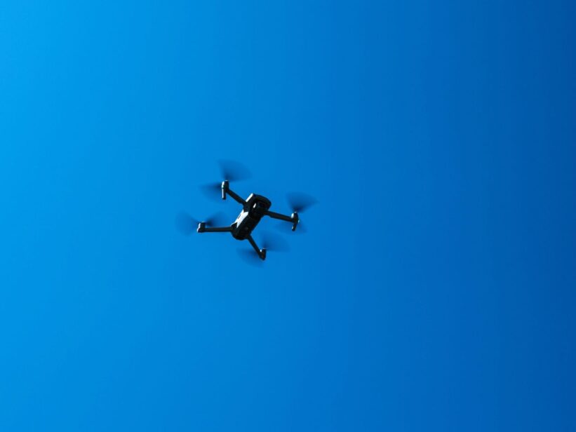 A black drone flying high in a clear blue sky, showcasing modern technology and flight capabilities.