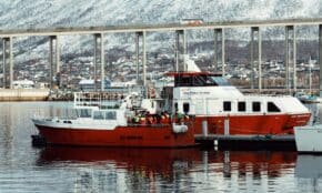 Red fishing boat docked in Tromsø, Norway with snow-covered mountains and a bridge in the background.