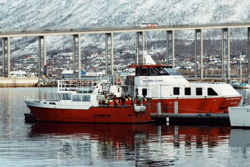 Red fishing boat docked in Tromsø, Norway with snow-covered mountains and a bridge in the background.