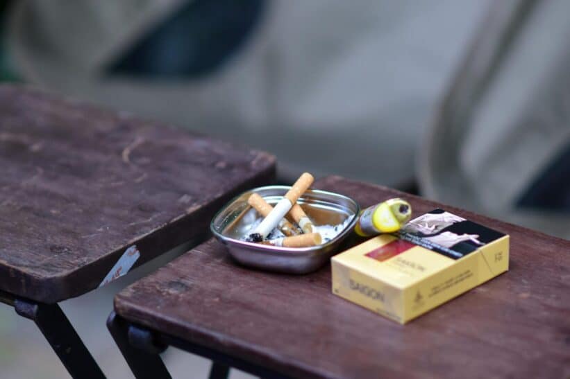 Close-up of ashtray with cigarette butts and a pack of cigarettes on a rustic wooden table.