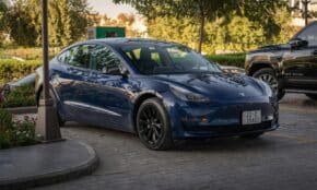 Electric blue Tesla Model 3 parked in sunlit commercial parking area.