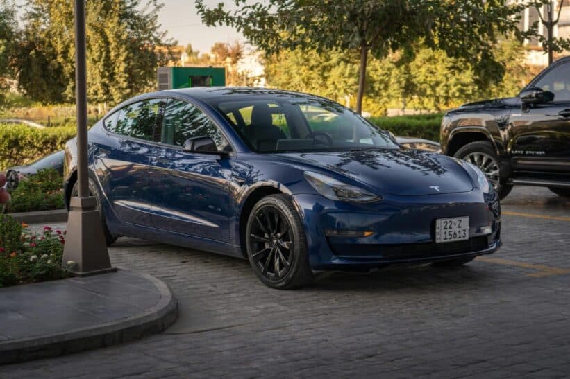 Electric blue Tesla Model 3 parked in sunlit commercial parking area.