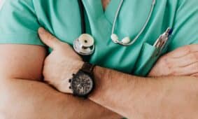 Unrecognizable female doctor in medical uniform with wristwatch on hand crossing arms on chest while standing with stethoscope on neck