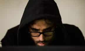 Focused young male hacker wearing black hood browsing dark net on netbook and hacking database in light room