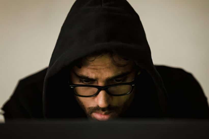 Focused young male hacker wearing black hood browsing dark net on netbook and hacking database in light room