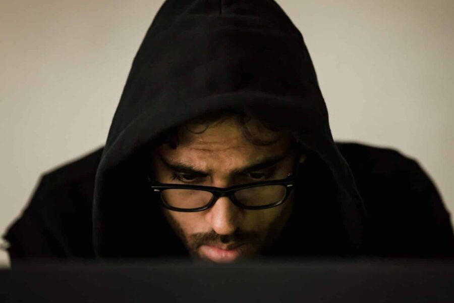 Focused young male hacker wearing black hood browsing dark net on netbook and hacking database in light room