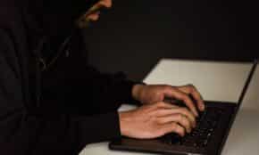 Crop bearded male hacker in black hood typing on netbook keyboard and browsing dark net in obscure room