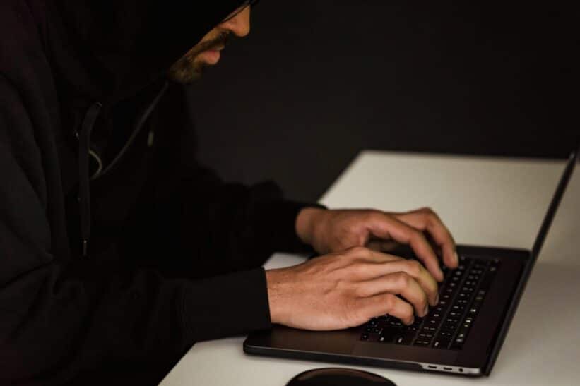 Crop bearded male hacker in black hood typing on netbook keyboard and browsing dark net in obscure room