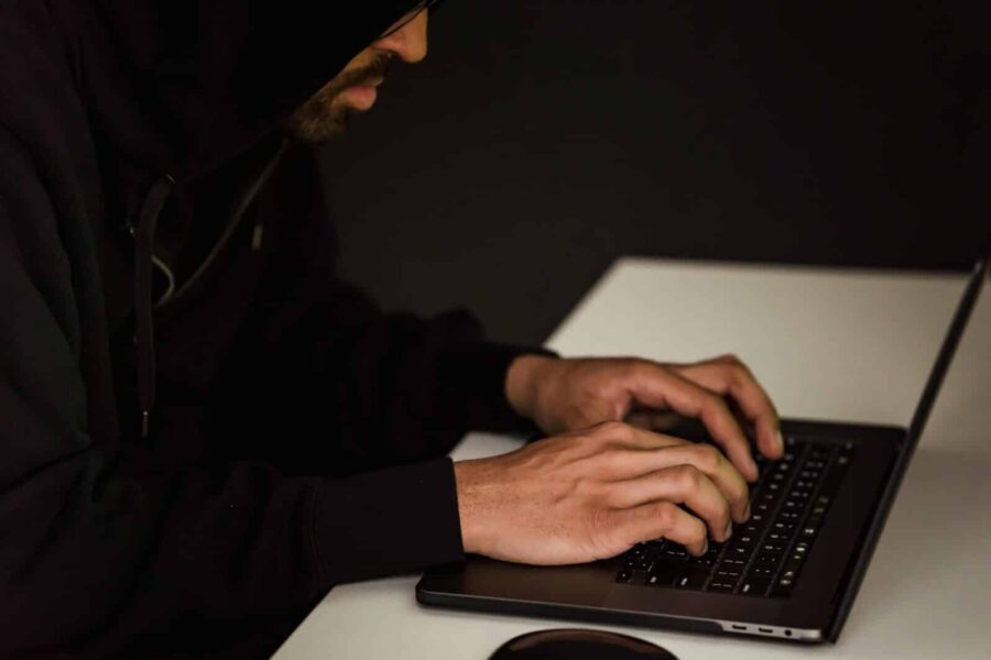 Crop bearded male hacker in black hood typing on netbook keyboard and browsing dark net in obscure room