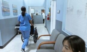 A nurse in scrubs pushes a wheelchair through a well-lit hospital corridor, attending to patient care.