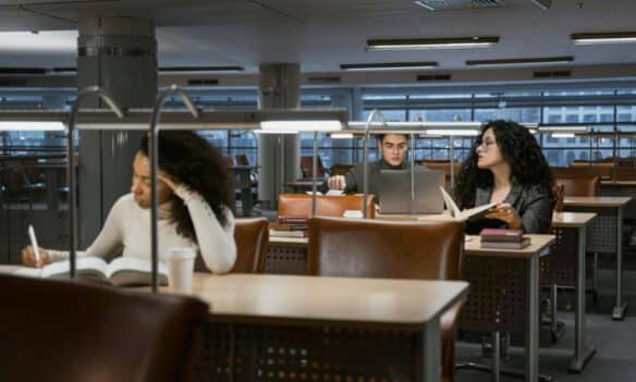 College students studying in a modern library setting, concentrating on books and laptops.