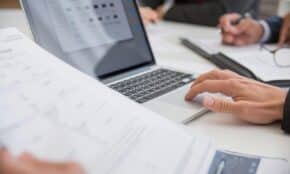 Close-up of hands working on documents and a laptop in an office setting, illustrating teamwork and productivity.