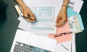 Hands writing on tax documents with laptop, glasses, and currency on desk.