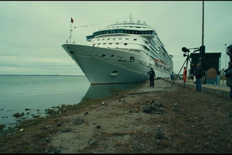 Cruise ship docked in Argentina with passengers disembarking as news crews film, marshy area with rodents visible nearby